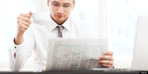 Handsome businessman reading newspaper with laptop and coffee cup in office.