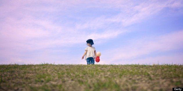 Toddler girl holding balloons on top of grassy hill against backdrop of blue sky.
