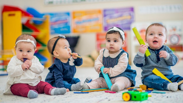Group of toddlers sitting indoors in a daycare center. They are playing with colorful wooden blocks. Some of the kids are putting the blocks in their mouths.