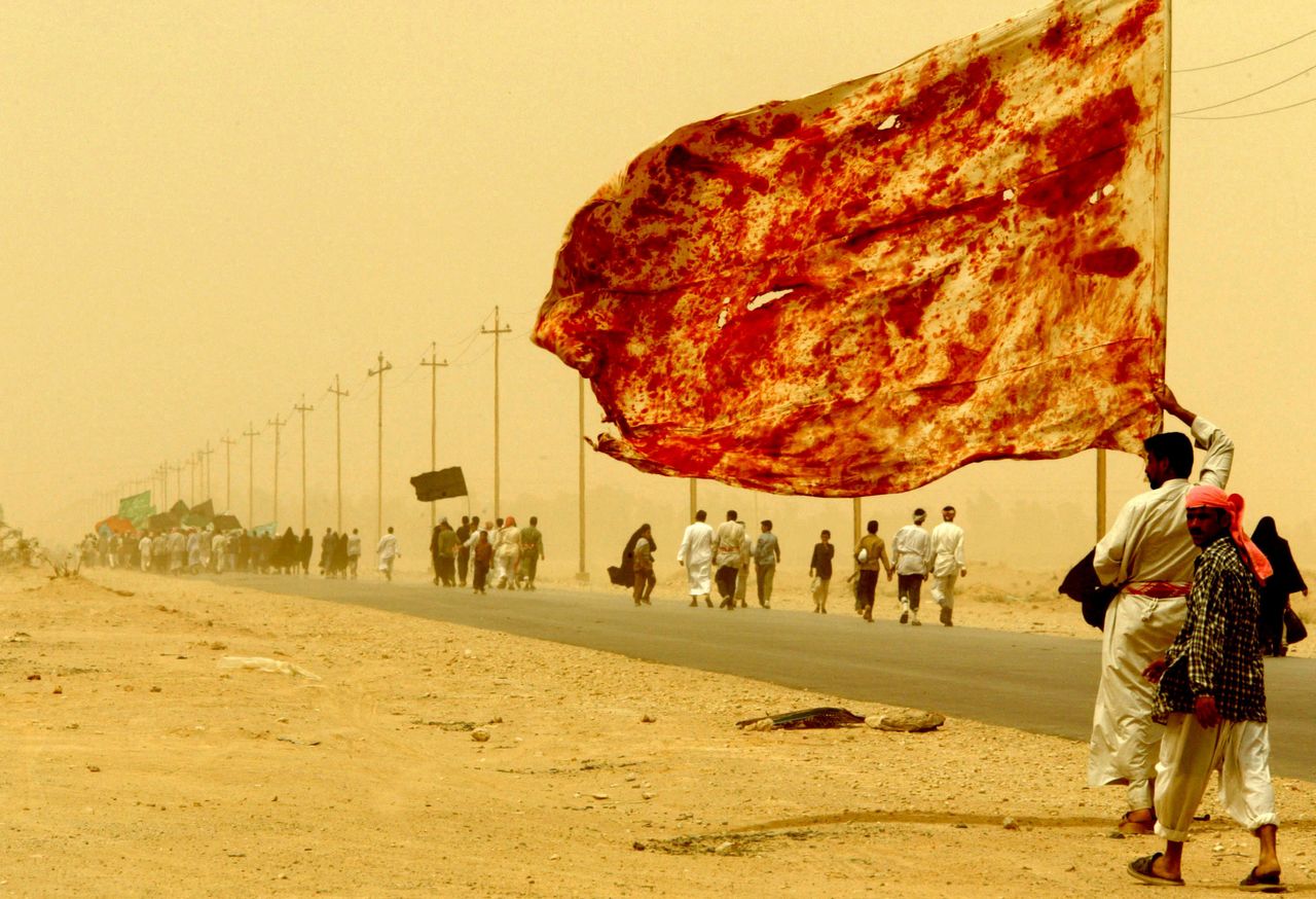Ιρακινοί περπατούν εν μέσω αμμοθύελλας προς την Κερμπάλα. Ιράκ, 19 Απριλίου 2003. Η σημαία που κρατά ο άνδρας είναι «βαμμένη» με αίμα σιιτών μουσουλμάνων.