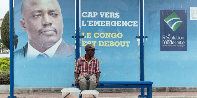 A vendor sits at a Kinshasa bus stop in front of a poster featuring the Democratic Republic of Congo president.