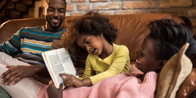 Happy African American parents enjoying with their little daughter at home while she is reading a book to them. Focus is on little girl.