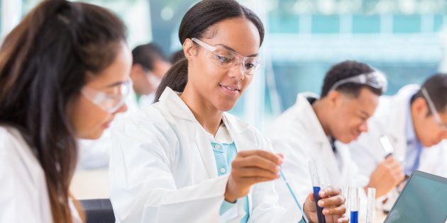 African American female chemist drops liquid into a beaker. Her assistant is watching her. She is recording the findings on a laptop.