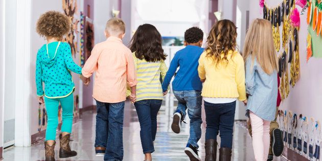 Rear view of a multi-ethnic group of seven children holding hands, walking down a school hallway together. They are in kindergarten or preschool 4 to 6 years old.