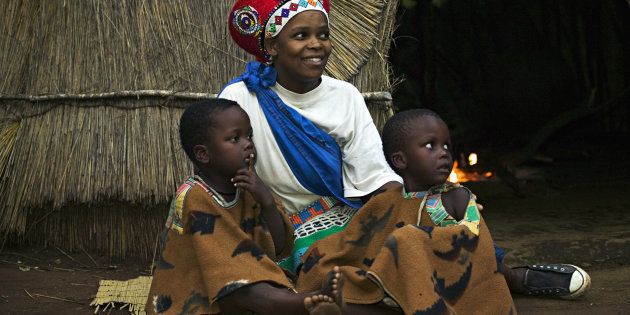 Zulu woman in traditional red headdress of a married woman with her children. Beehive hut in the background. Lesedi Cultural Village near Johannesburg, South Africa.