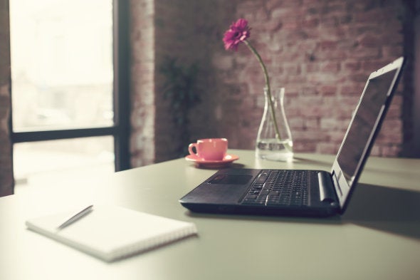 Close-up shot of a working place: laptop computer, notebook with pencil, espresso coffee and gerbera daisy flower in vase on a work desk.