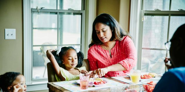 Mother cutting young daughters food while eating breakfast together at dining room table