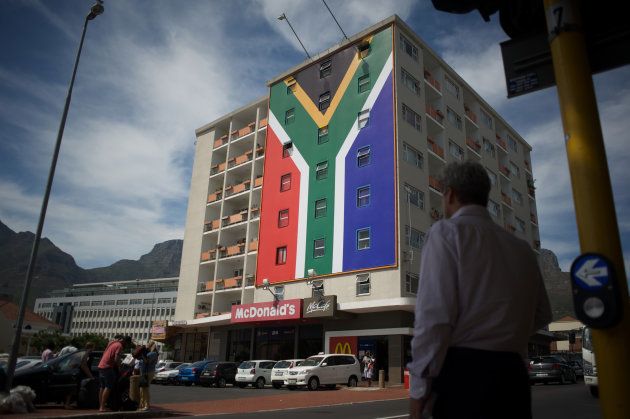People walk past the billboard of the South African flag, newly set up on the building in the trendy Long Street area, where a billboard saying 'Zuma must fall' was set up some days earlier, and then destroyed, on January 22 2016, in Cape Town. The original billboard referred to South African president, and African National Congress(ANC) president, Jacob Zuma.