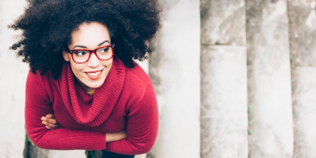 Portrait of smiling young woman standing arm crossed on staircase. High angle view. Woman with curly hair, red eyeglasses, and red pullover.