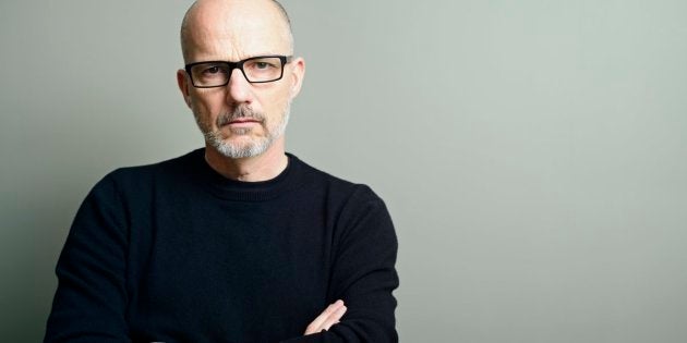 A handsome, balding mature man with arms crossed, wearing black-rimmed glasses, black t-shirt and sweater, looks seriously at the camera.