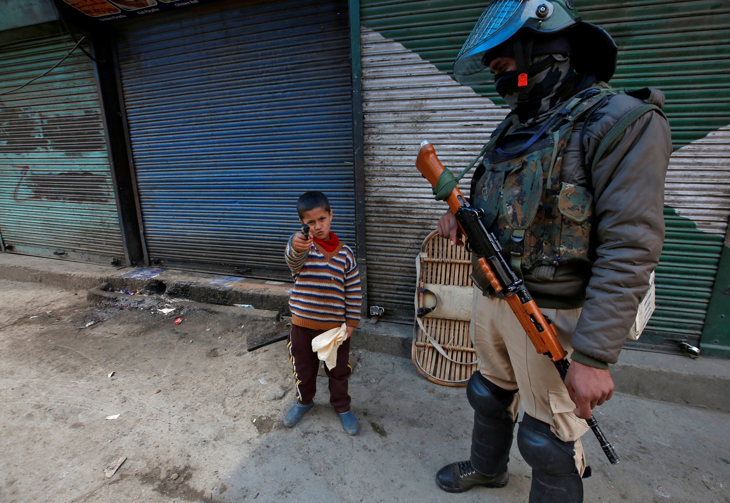 A boy plays with his toy pistol next to an Indian policeman standing guard in front of closed shops during a strike called by