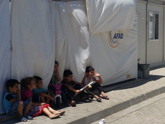 Varios niños ríen bajo la fina sombra proyectada por un contenedor vivienda. | Campo de Kahramanmaraş.