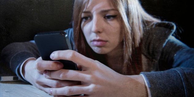 young scared teenager girl looking worried and desperate to mobile phone as internet stalked victim abused and cyberbullying or cyber bullying stress concept in black background
