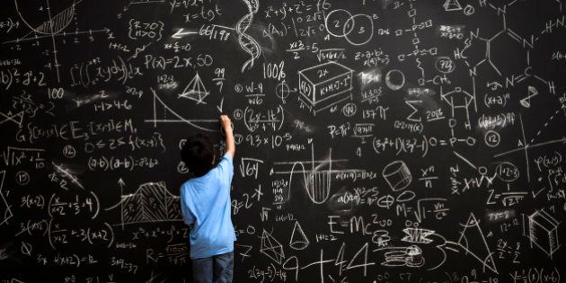 A young boy stands drawing on a huge chalkboard filled with mathematical equations