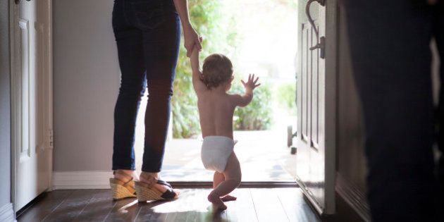 Rear view of mother holding baby boys hands standing at open front door