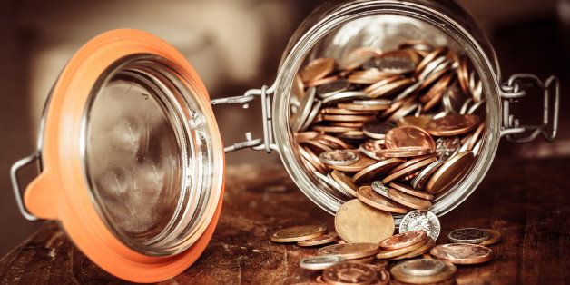 A jar of sterling coins is tipped on its side with the coins spilling out on to a wooden surface. The jar denotes a savings pot.