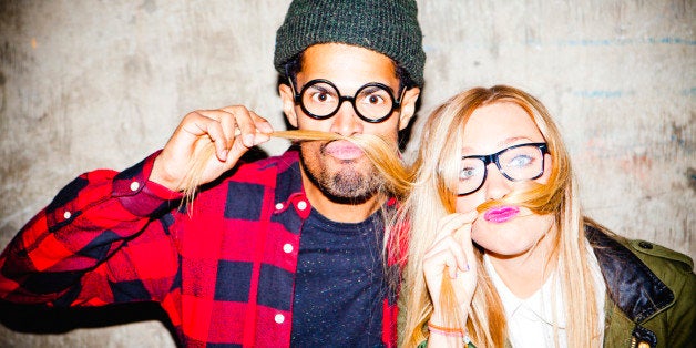 Portrait of a young man and woman pulling funny faces and messing around, standing in front of a rustic background.