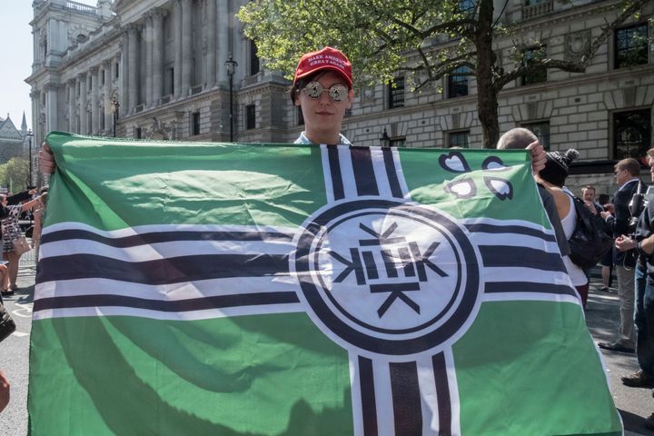 A man at a Tommy Robinson rally in London holds an alt-right "Kekistan" flag while wearing a Make America Great Again hat.