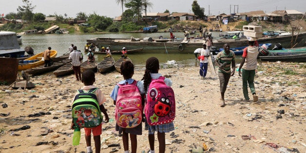 Three schoolchildren, carrying brightly colored backpacks, stand and wait for a canoe to transport them across the Nun River