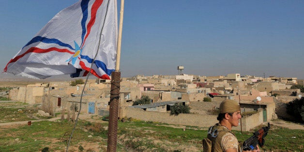 A Dwekh Nawsha militia member stands next to a flag of the Assyrian Patriotic Party, as he stands guard on the rooftop of a building in the Christian village of Bakufa, 30 kilometers (18.6 miles) north of Mosul, Iraq, Wednesday, Nov. 12, 2014. The party flag has replaced the black flag of the Sunni militants of the Islamic State group and is waving on the roof of the building at the entrance to the village. (AP Photo/Bram Janssen)