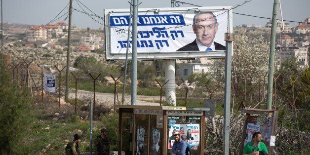 Israeli soldiers (L) stand guard next to settlers at a bus stop next to campaign posters of two candidates running for general elections: Israeli Prime Minister and Likud party's Benjamin Netanyahu (top) and extreme right-wing activist Baruch Marzel on March 10, 2015 in the Jewish settlement of Ofra in the West Bank, north of Jerusalem. AFP PHOTO / MENAHEM KAHANA (Photo credit should read MENAHEM KAHANA/AFP/Getty Images)