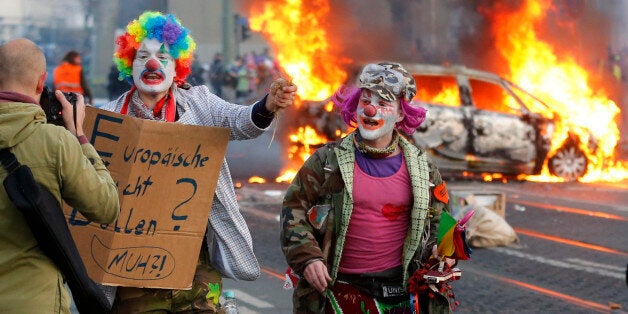 Demonstrators dressed as clowns pass by a burning police car Wednesday, March 18, 2015 in Frankfurt, Germany. The Blockupy alliance said activists plan to try to blockade the new headquarters of the ECB to protest against government austerity and capitalism. (AP Photo/Michael Probst)