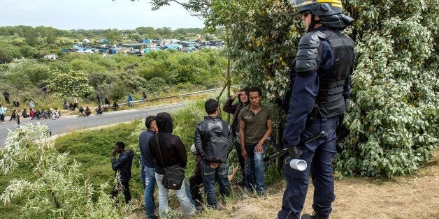 A French riot police officer (CRS) stands in front of men on the side of the road as illegal migrants attempt to hide in lorr
