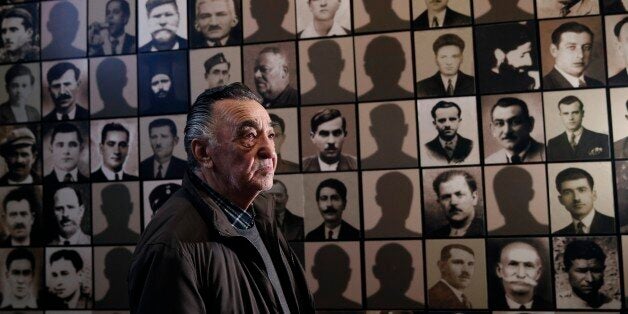 In this photo taken Saturday, March 21, 2015, Georgios Dimopoulos, 85, a survivor of the Kalavryta massacre poses for a photograph next to the portraits of his father (fourth row, fifth column) and his uncle (fifth row, third column) at the Holocaust Museum in the town of Kalavryta ,western Greece. On December 13, 1943, more than 500 males aged 14 and over were machine-gunned in the greatest massacre perpetrated by the occupying German troops. Greek Prime Minister Alexis Tsipras, who will meet Chancellor Angela Merkel in Berlin Monday, has revived the issue of German compensation for crimes like these. The Germans have responded that the issue of reparations has been conclusively settled. (AP Photo/Petros Giannakouris)