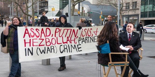 UNITED NATIONS, FIRST AVENUE, NEW YORK, NY, UNITED STATES - 2015/12/18: Taking advantage of the heightened media presence at the United Nations, demonstrators rally behind TV anchors as they record an interview. Around the same time that the ministerial International Syrian Support Group meeting in New York City drew to a close, and its proposal for a ceasefire agreement regarding Syria was brought to the United Nations Security Council; several dozen activists rallied across from UN Headquarters, condemning Russia's support for Bashar Al-Assad's government. (Photo by Albin Lohr-Jones/Pacific Press/LightRocket via Getty Images)