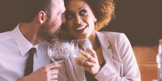 Happy couple - afro american woman and caucasian man in elegant outfits having lunch or dinner in restaurant, sitting at the table and holding wine glasses. Man embracing her girlfriend.
