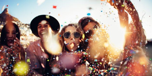 Group of teenager hipster friends partying by blowing and throwing colorful confetti from hands with sunset sun flare