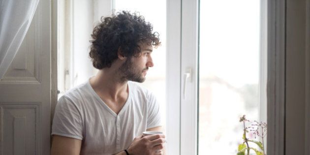 Young serious man at home thinking while holding cup of coffee and looking through the window