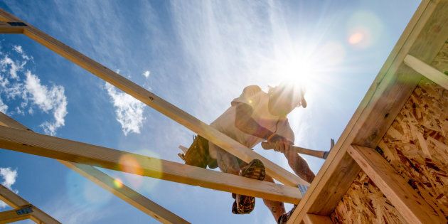 Manual laborer poised on a new construction hammering a piece of frame work with The sun and a partly cloudy blue sky in the background.
