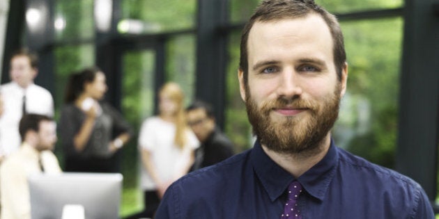 Portrait of young business man. He is wearing a blue shirt and dotted purple tie. There are five business associates in the background. He is in a glass building that is surrounded by trees.