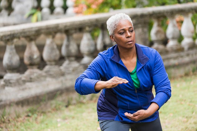 A senior African American woman in the park, practicing Tai Chi. She has a serious expression on her face as she concentrates on her pose. She is wearing a royal blue jacket over a green shirt and gray leggings.