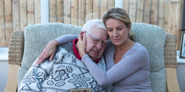 A shot of a senior man sitting on the sofa with his daughter, looking at a photograph. He is cuddling his daughter, looking sad and holding the photo.