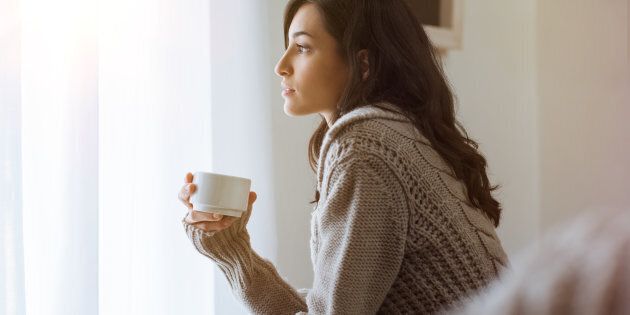 Young woman looking over window pane holding coffee. Thoughtful woman thinking and looking away while drinking hot tea. Woman in warm sweater looking outside window while drinking tea at morning.