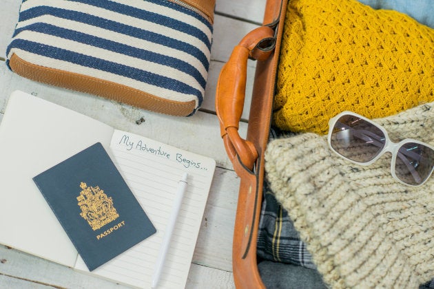 A series of travel items are prepared and laid out on a white wooden background. Included amongst these items are a notebook, wallet, purse, leather suitcase, passport, pen, sunglasses, and clothing. The items are ready for a vacation.