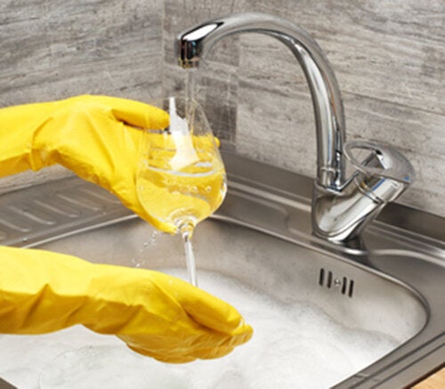 80674783 - close up of female hands in yellow protective rubber gloves washing wine glass under running tap water against kitchen sink full of foam and tableware