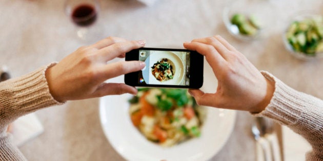 Bird's eye view of a woman taking a photo of her dinner with her smartphone. The plate is seen through the screen of the phone, while there is more food, cutlery, wine and bread on the table.