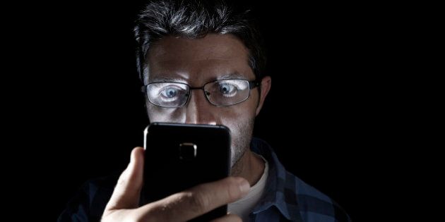 close up portrait of young man looking intensively to mobile phone screen with blue eyes wide open isolated on black background on dark edgy lighting scheme in addiction to internet technology