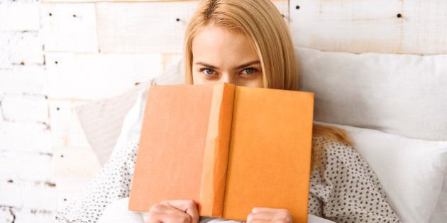 Reading enjoyment. Carefree young woman is holding book and covering her face with it. She is sitting in bed and looking forward with joy