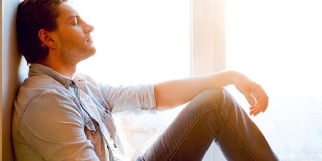 Side view of handsome young man sitting at the window sill and keeping eyes closed