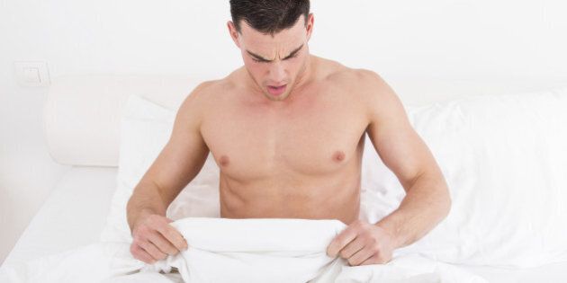 surprised half naked young man in bed looking down at his underwear at his penis under white covers sheet in badroom. Concept photo of male sexuality and man sex problems, domestic atmosphere.