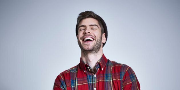 Portrait of young bearded man with tattooed arms in red and blue check shirt and beanie, laughing with head back and eyes shut