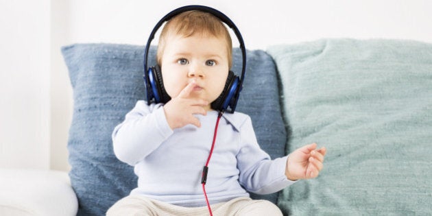 Sweet baby boy sitting on sofa and listening music at headphones while holding one finger in front of his mouth.