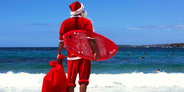 Man dressed as Santa Claus holding sack and surf board standing on beach, rear view