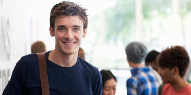 Portrait of smiling university student standing in corridor during break, people in background talking