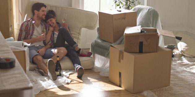 A young professional couple sitting on sofa cushions in their new house surrounded by packing boxes. they are drinking champagne.