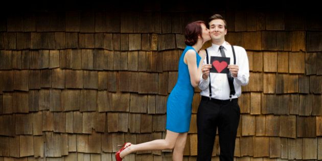 Young urban styled couple. Man poses grinning with a heart shaped valentine while girl kisses him on the cheek with one leg kicked up in the air.